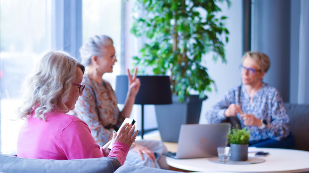 Etualalla vaaleanpunaiseen paitaan pukeutunut harmaatukkainen nainen selaa kännykkää ja taustalla kaksi naista keskustelee digitaalisuudesta. Pöydällä avoin läppäri.