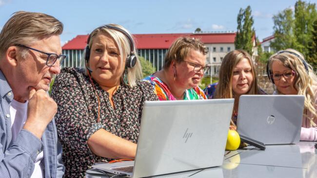 Ihmisjoukko ulkona pöydän ympärillä. Kahdella on kannettavat tietokoneet, kaikki keskustelevat keskenään. Kesäinen ilma.