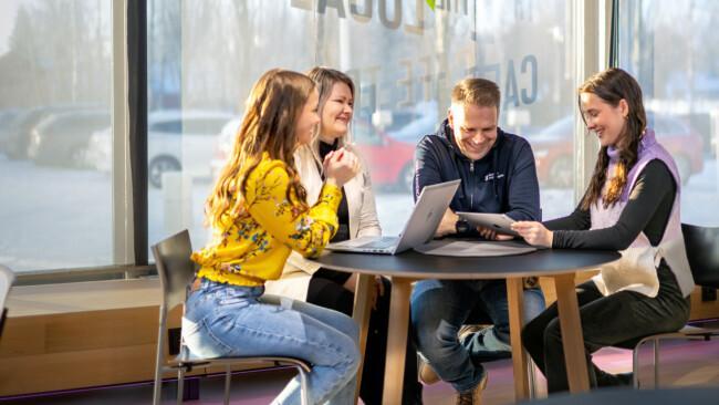 Työpaikkaohjaaja työssään. Neljä henkilöä pöydän ympärillä iloiset ilmeet kasvoillaan perehtyvät työpaikan ja opintojen asioihin.
