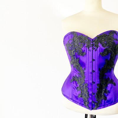 Violetti sovitusnuken päällä oleva rinnanylikorsetti, jossa mustia koukeroisia koristeita.