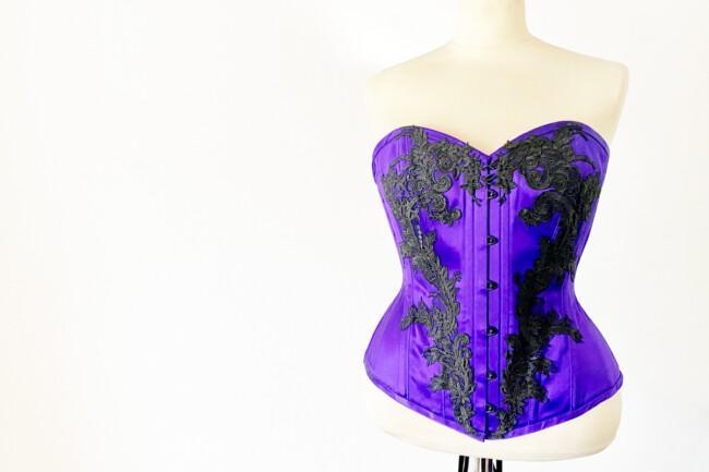Violetti sovitusnuken päällä oleva rinnanylikorsetti, jossa mustia koukeroisia koristeita.