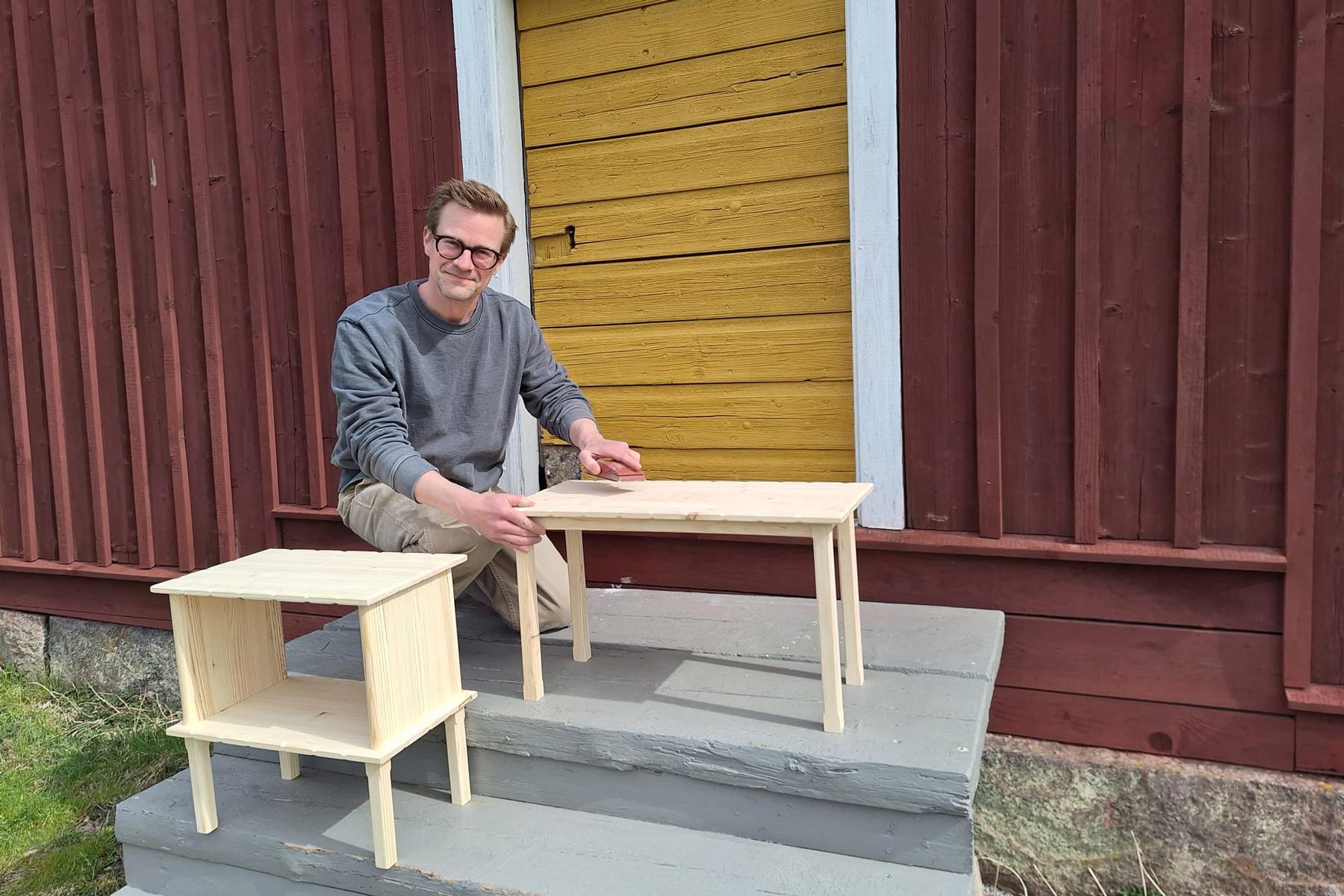 Mies valmistamiensa puuhuonekalujen vieressä.